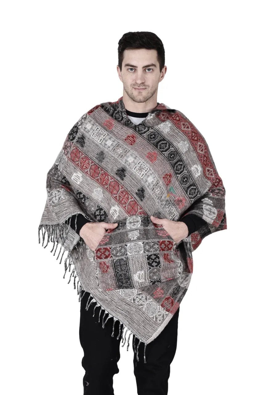 A man stands facing forward with his hands in the front pocket, wearing an Aruna Kullu Handloom grey kullu shawl woolen poncho with hood, featuring red, black, and gray designs, against a plain white background.