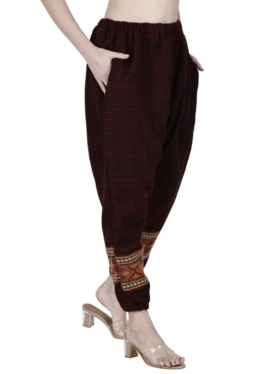 A person wearing loose yoga pajama maroon singal patti with ethnic patterns near the ankles, standing with one hand in a pocket and clear heeled sandals. Only the lower half of the body is visible.