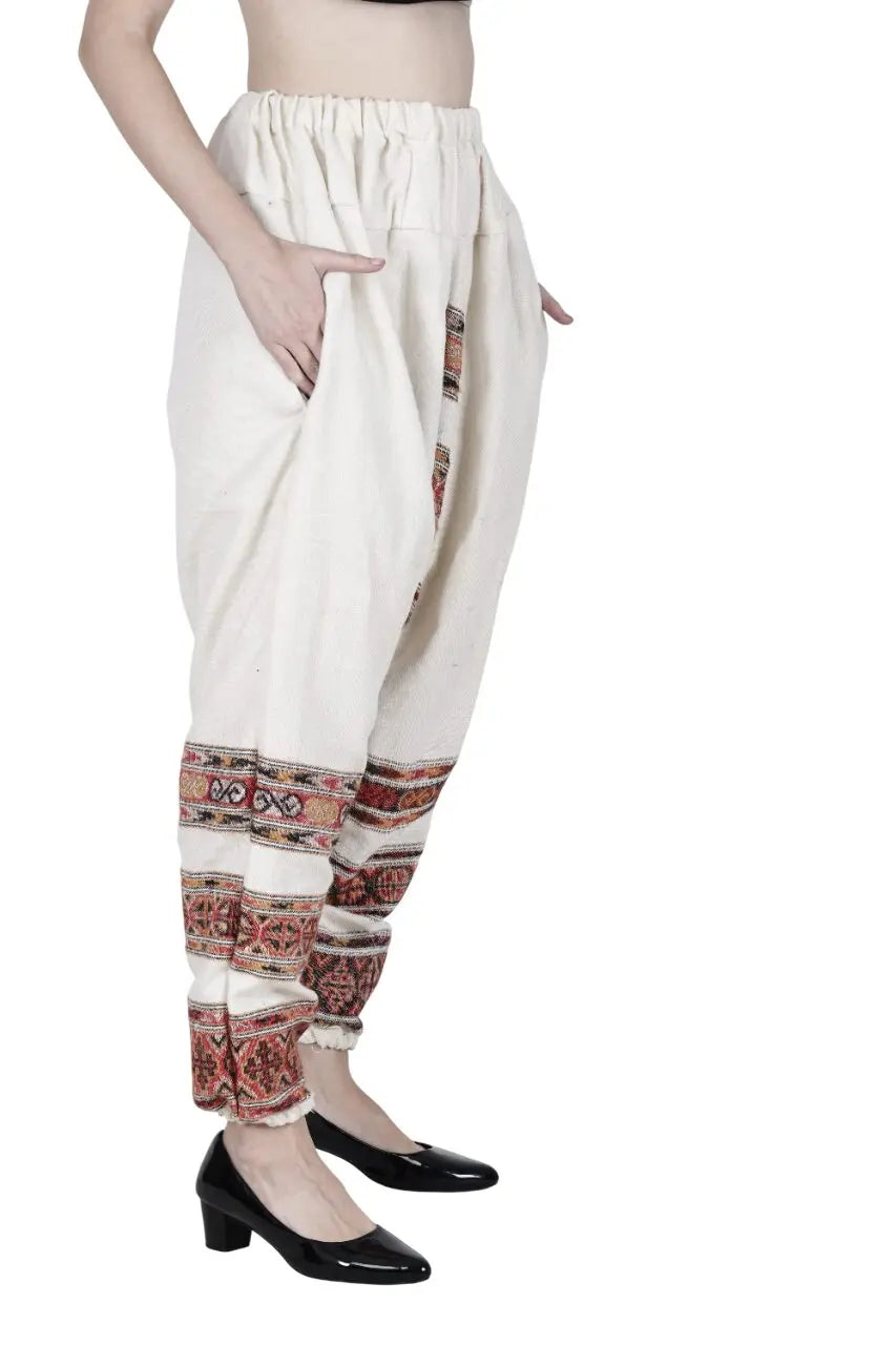 A person wearing high-waisted, loose yoga pant white 3 patti from aruna kullu handloom, featuring colorful embroidery near the ankles and paired with black patent heels. Photo shows only the lower torso and legs from the side.