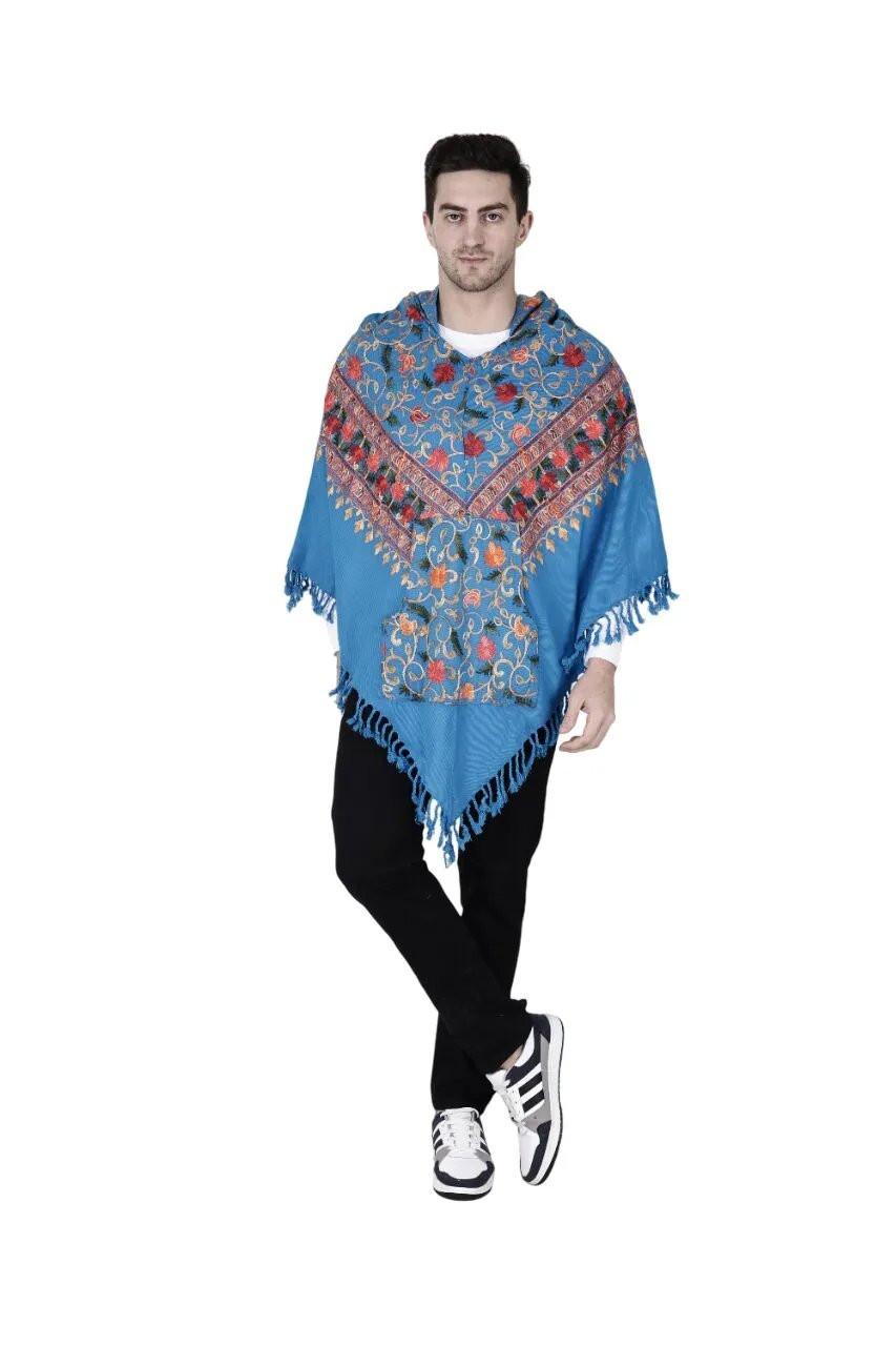 A man wears the Aruna Kullu Handloom Kullu Shawl Woolen Poncho with Hood Kashmiri Blue, featuring colorful handwoven floral embroidery and fringe edges, paired with black pants and white sneakers, standing against a white background.