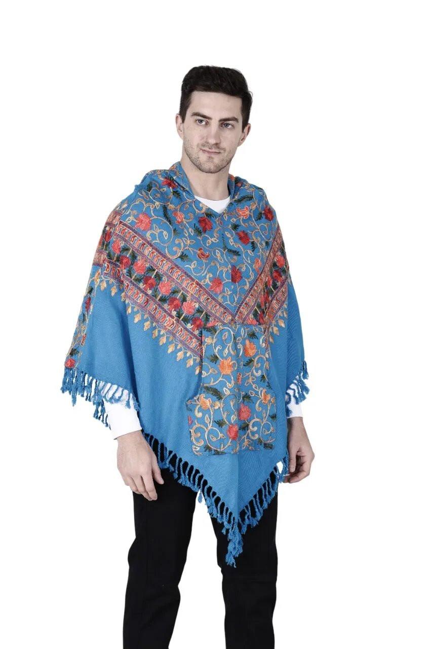 A man wears the "kullu shawl woolen poncho with hood kashmiri blue" by aruna kullu handloom, featuring colorful floral and geometric patterns with fringed edges, over a white shirt and black pants, set against a white background.