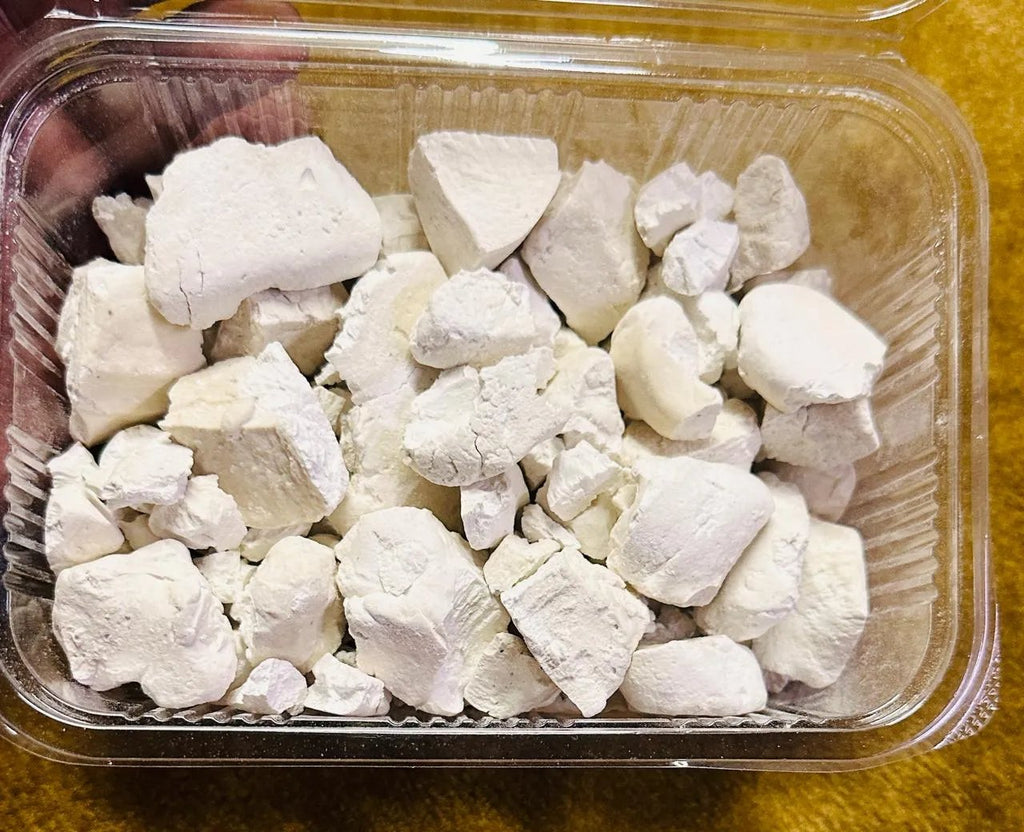 A 500GM plastic container of Himachali Sheera by Aruna Kullu Handloom, filled with irregular white chalky chunks, rests on a yellow surface.