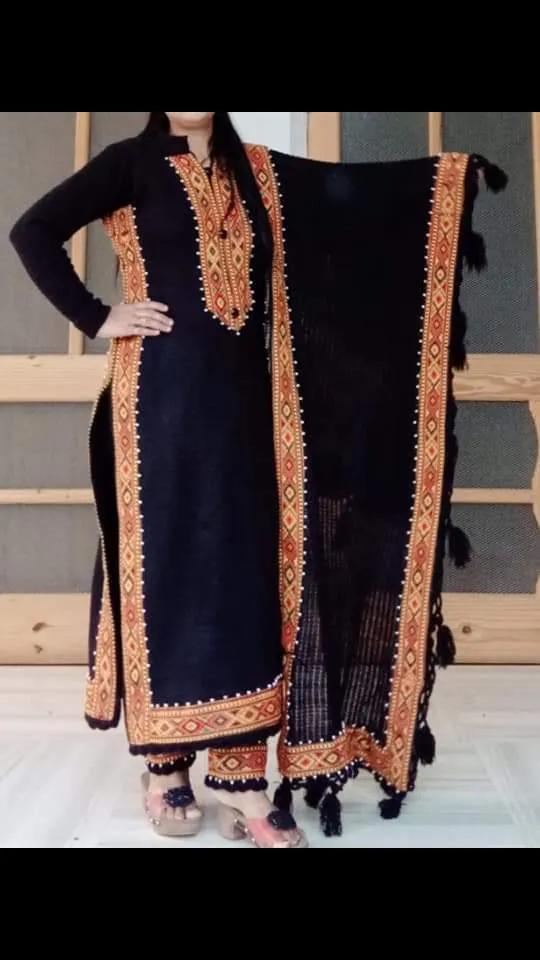 A woman wears a suit adorned with aruna kullu handloom’s KULLU Patti (LACE) Border for Suits Kurtis and other dress material (3 M), paired with matching pants, a tasseled dupatta, and Pula Chappal sandals, standing indoors by wood and glass.