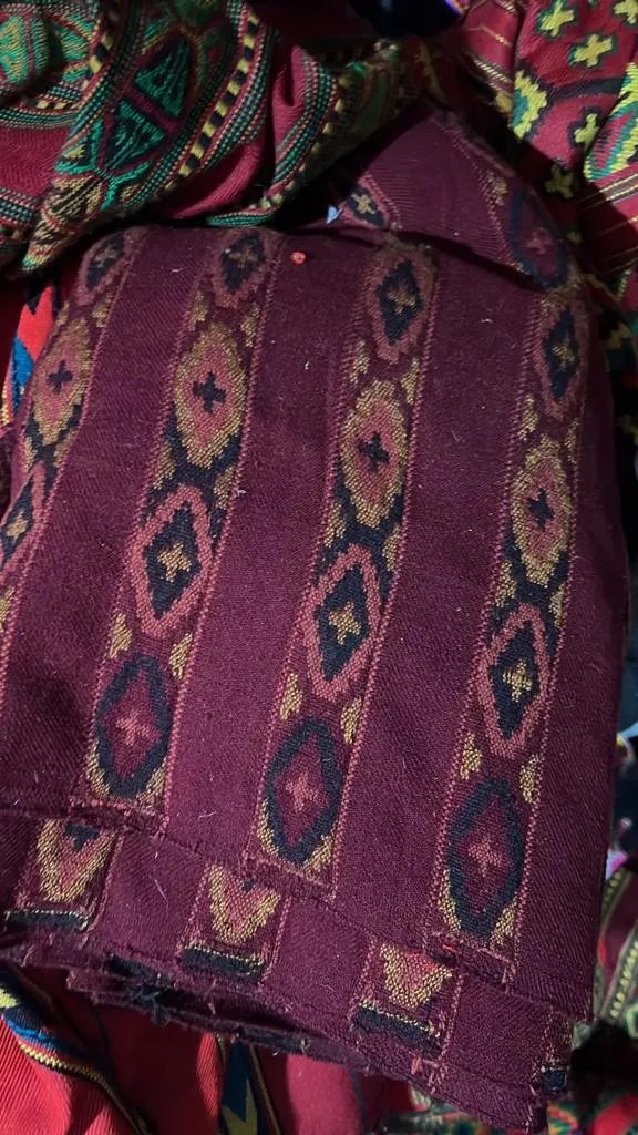A close-up of aruna kullu handloom’s KULLU Patti (LACE) Border for Suits, Kurtis, and Dress Material (3 M) shows maroon fabric with black, gold, and red geometric patterns and a kullu border among other vibrant textiles.
