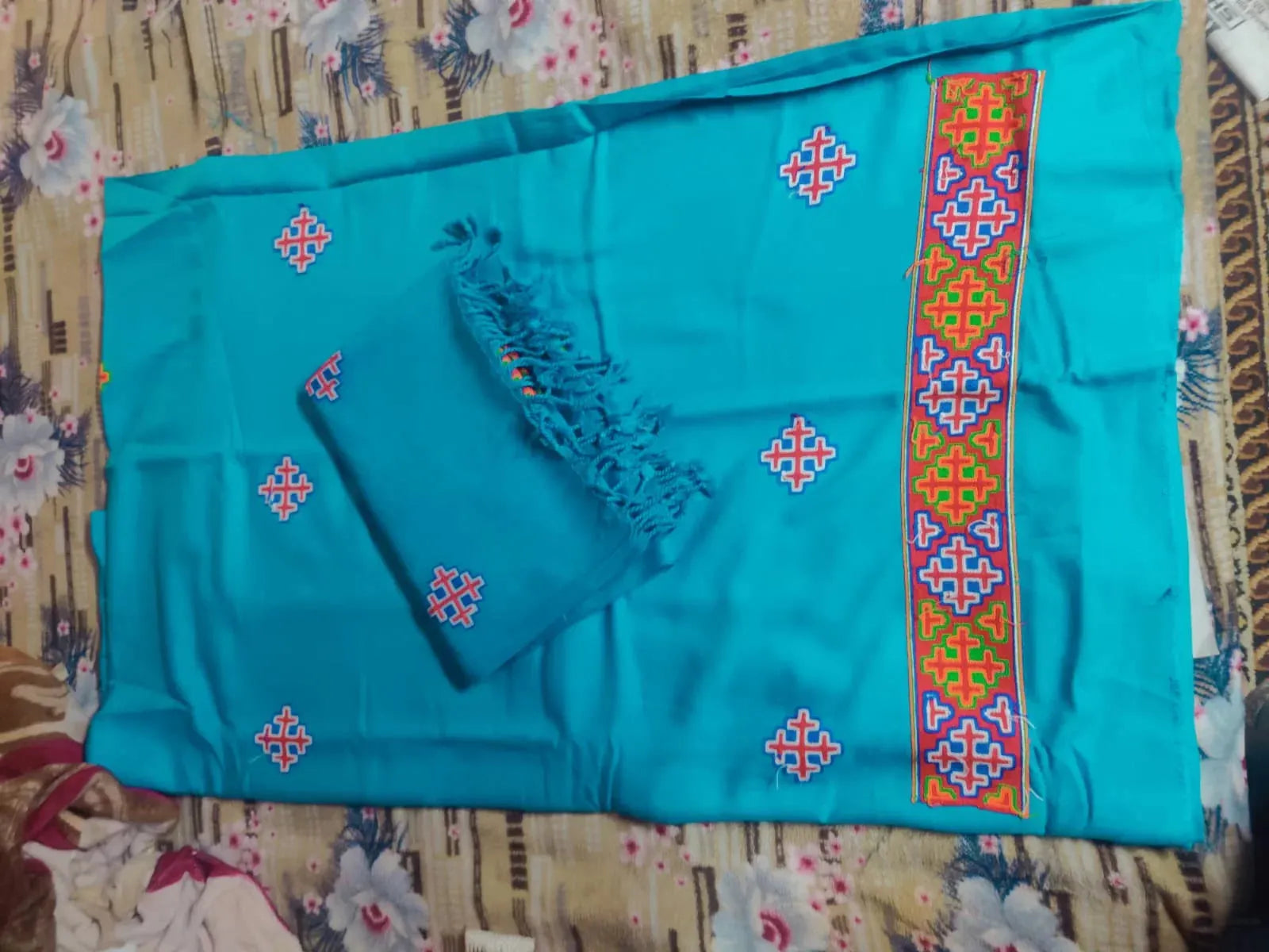 The aruna kullu handloom kullu patti woolen suit (4 meters) with frozen blue stole features colorful embroidered geometric patterns and a traditional border, styled with tassels, and is displayed on a floral-patterned bedsheet.