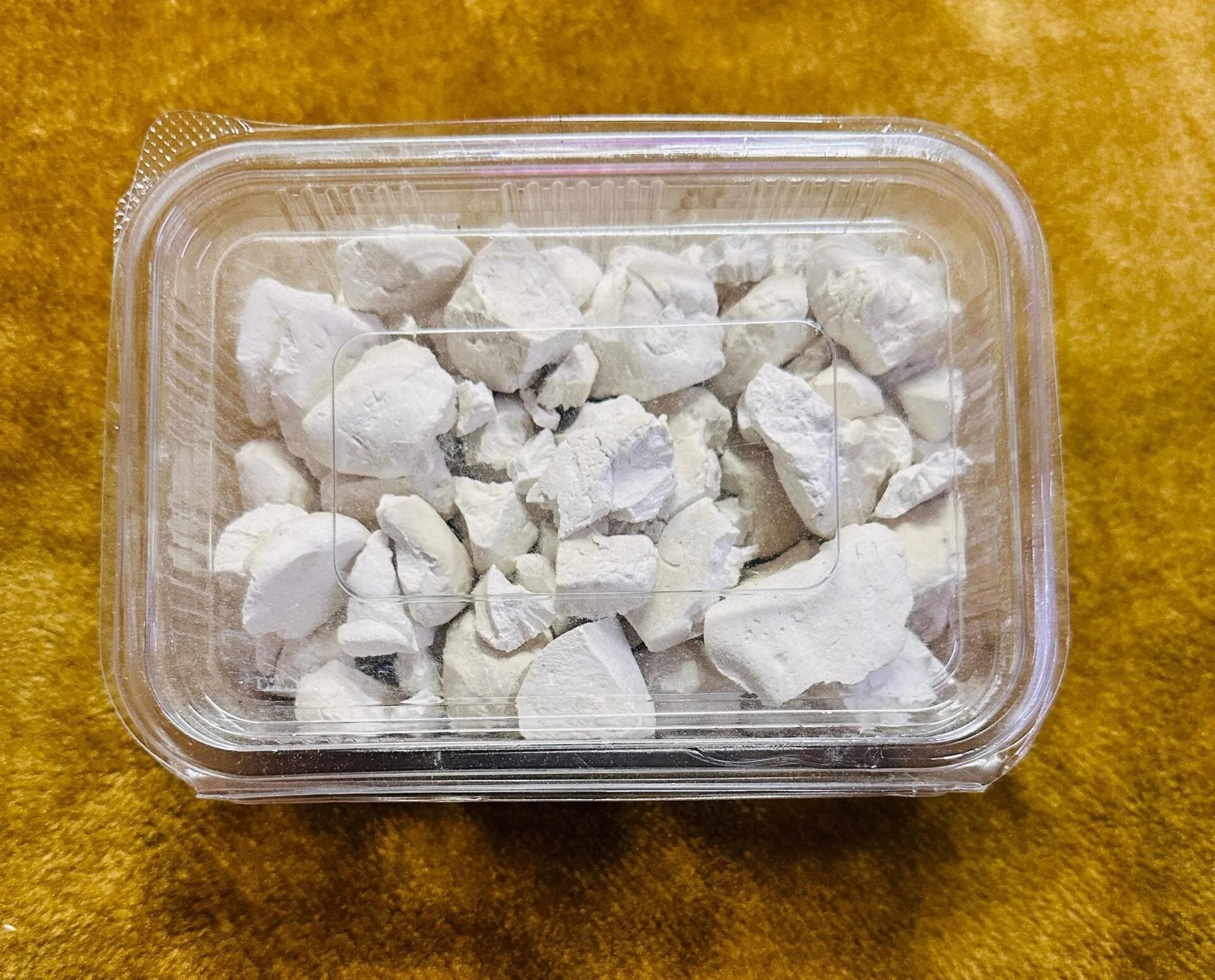 A clear plastic container of aruna kullu handloom himachali sheera 500GM, filled with irregular white chalky chunks, sits on a yellowish-brown textured surface.