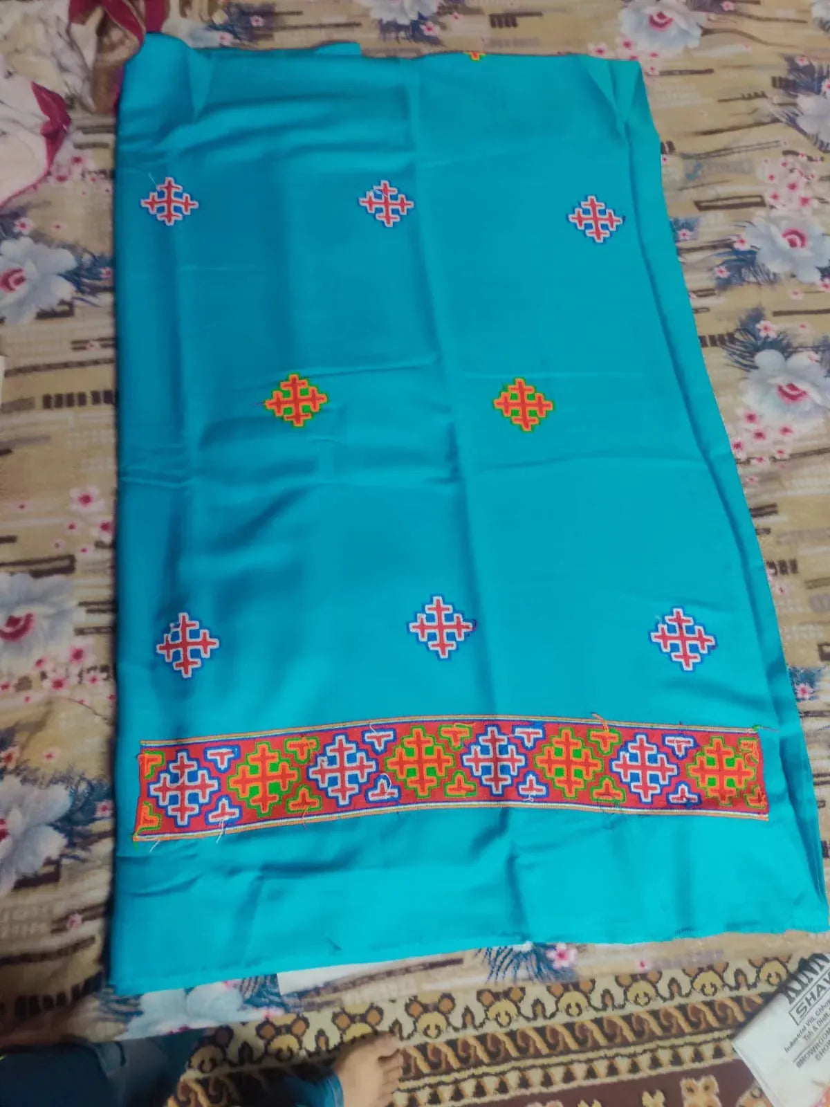 The aruna kullu handloom kullu patti woolen suit (4 meters) with stole in frozen turquoise features colorful geometric embroidery and a wide kullu patti border, displayed on a floral-patterned bedspread.