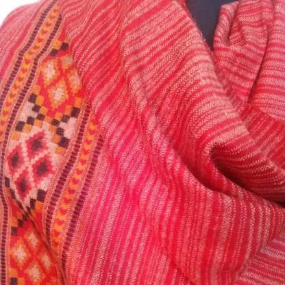 A close-up of the Kullu Kinnauri Handcrafted Shawl by aruna kullu handloom, featuring red woven fabric with yellow, orange, and black geometric patterns—stripes and diamond shapes—along one edge, draped in soft folds.