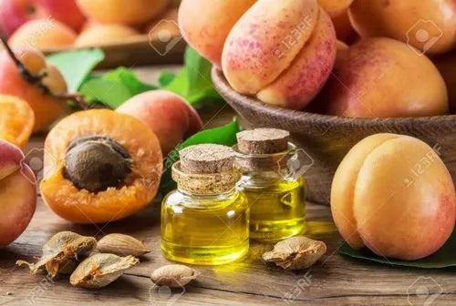 Close-up of apricots, seeds, and two small glass bottles of aruna kullu handloom GUTTI KA TEL (Apricot oil) on a wooden table. In the background, a bowl holds whole apricots and one fruit is cut open to show its pit.