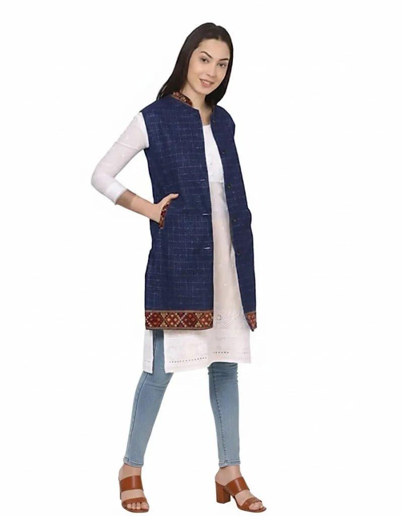 A woman smiles, wearing a long white kurta, aruna kullu handloom's check navy blue long jacket with kullu lace work, blue jeans, and brown heeled sandals, posing against a plain white background.half jacket for women khadi half jacket for ladies ladies half jacket cotton women's half jacket ladies sadri design winter half jacket for women
