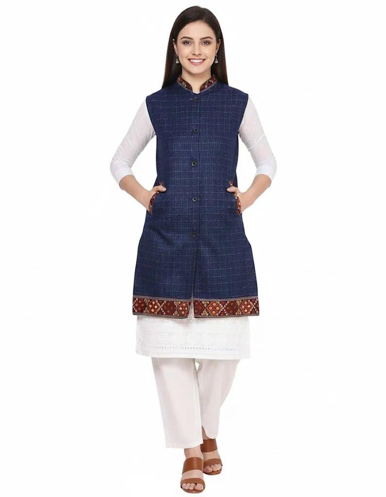 A woman smiles, posing in an aruna kullu handloom "Check navy blue long jacket with kullu lace work" over a white kurta and pants. She wears brown sandals and keeps her hands in the jacket's pockets against a white background.half jacket for women khadi half jacket for ladies ladies half jacket cotton women's half jacket ladies sadri design winter half jacket for women
